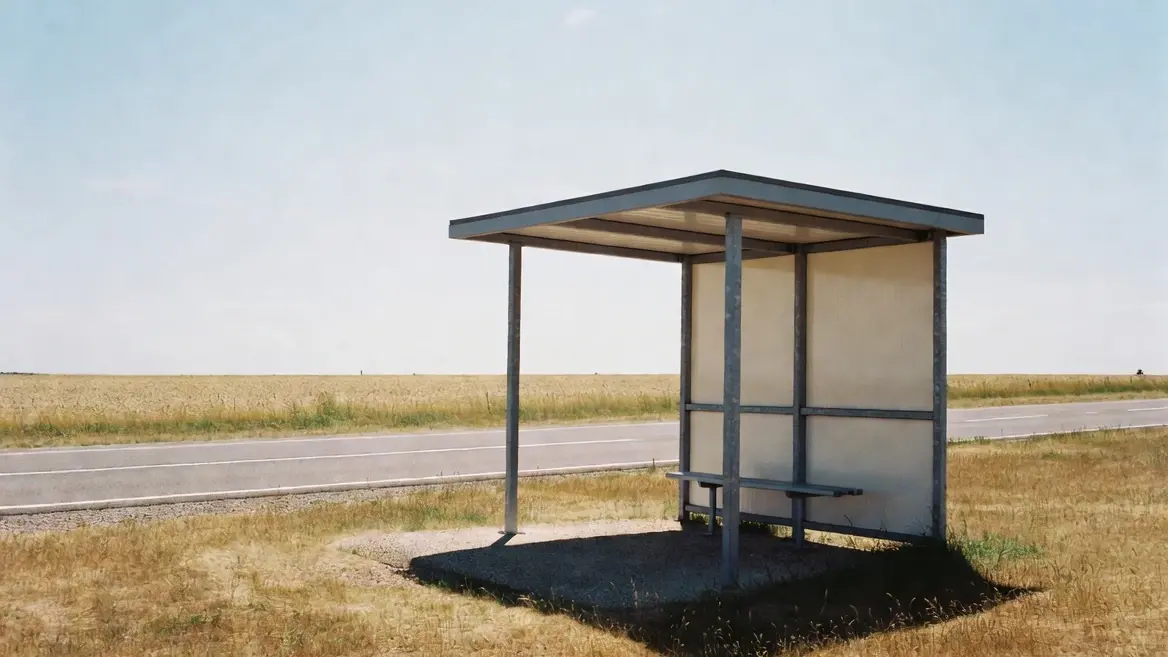 Eine leere Bushaltestelle mit flachem Metalldach steht an einer Landstraße zwischen trockenem Gras und Feldern.