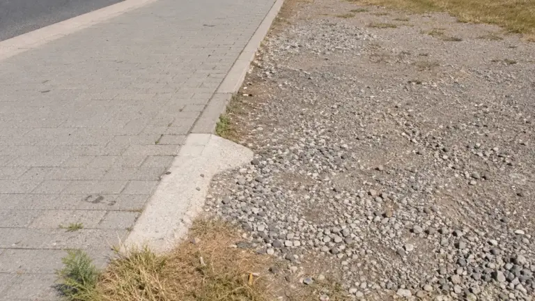 Eine abgesenkte Bordsteinkante verbindet einen gepflasterten Gehweg mit einer groben Schotterfläche an einer stillen Straße.
