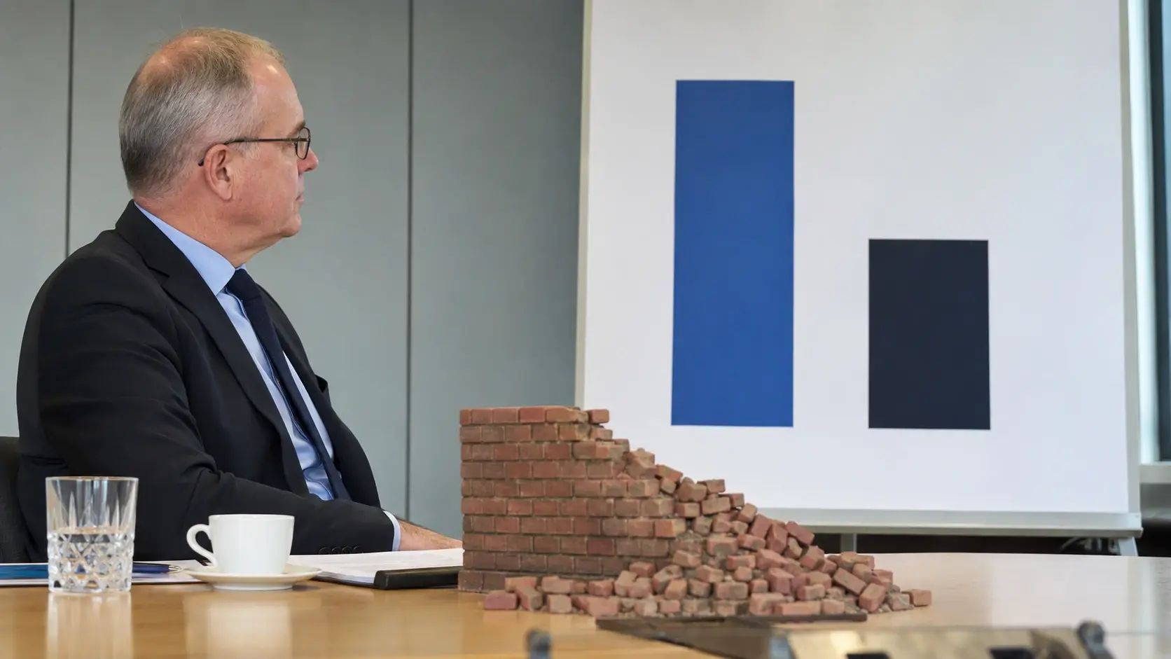 Ein älterer Mann im Anzug sitzt an einem Konferenztisch und blickt auf eine Tafel mit zwei Balken, während vor ihm ein teilweise eingestürztes Modell aus Ziegelsteinen auf dem Tisch liegt.