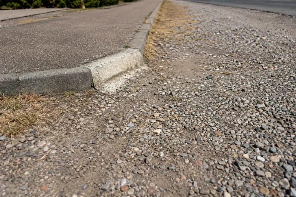 Aus niedriger Perspektive führt eine Bordsteinabsenkung von einem befestigten Weg in losen Kies und unebenen Untergrund.